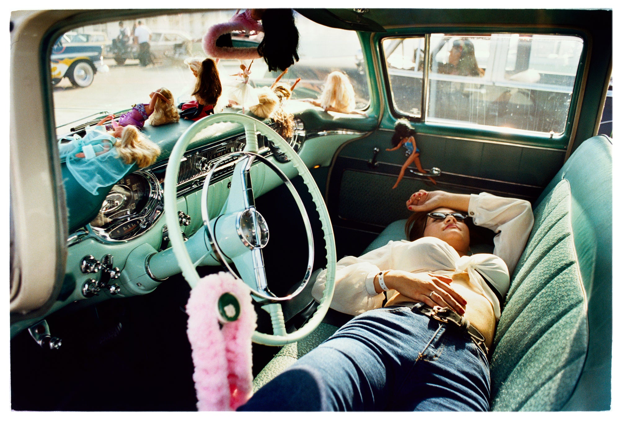 Photograph by Richard Heeps.  Inside a classic American car with a green interior, and dolls adorning the dashboard.  Lying on the seats is a woman resting.