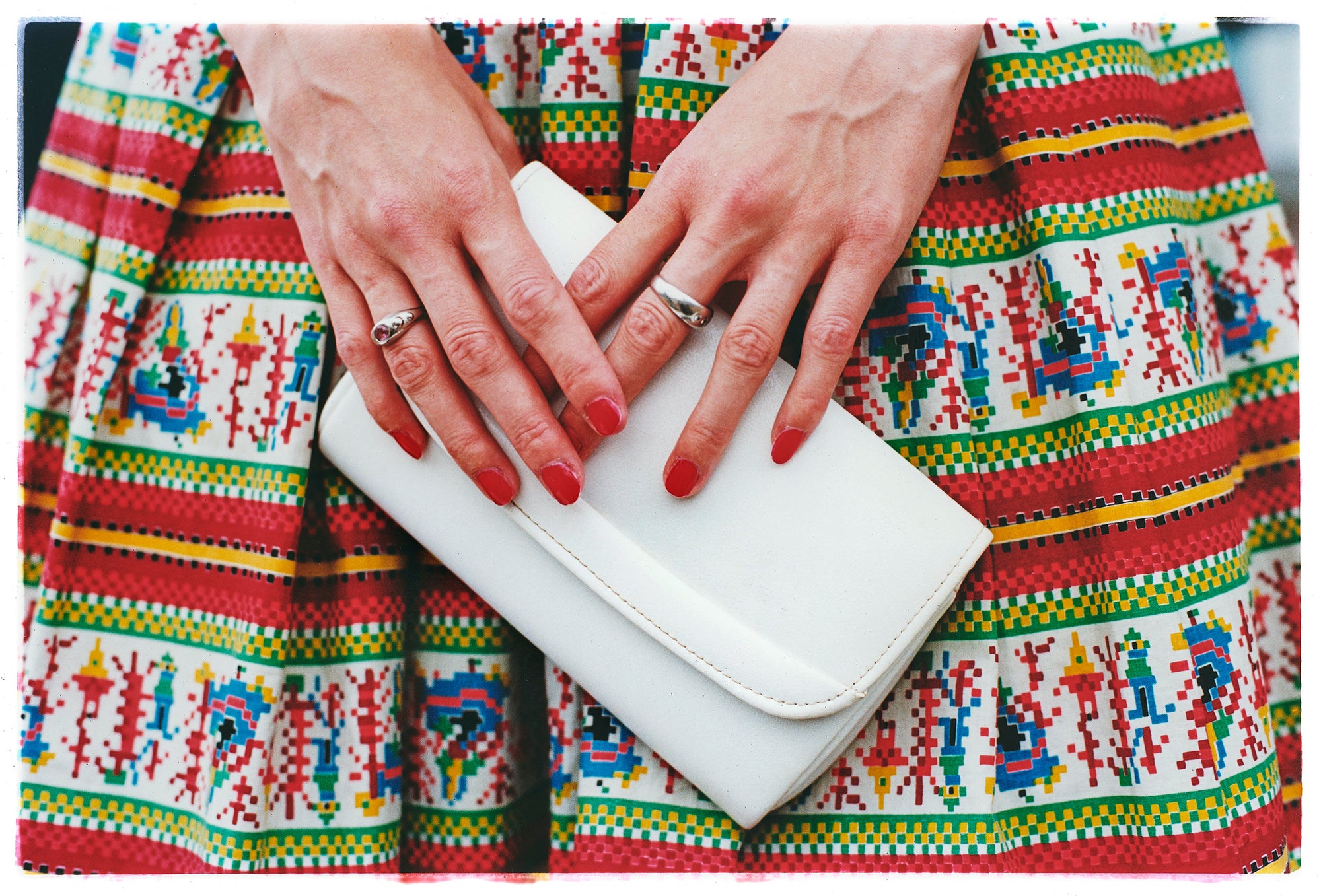 Photograph by Richard Heeps.  A white clutchbag, held by a pair of hands set against a vintage red patterned skirt.  