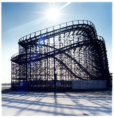 A rollercoaster structure throwing a shadow across the beach with the sun in a blue sky. 